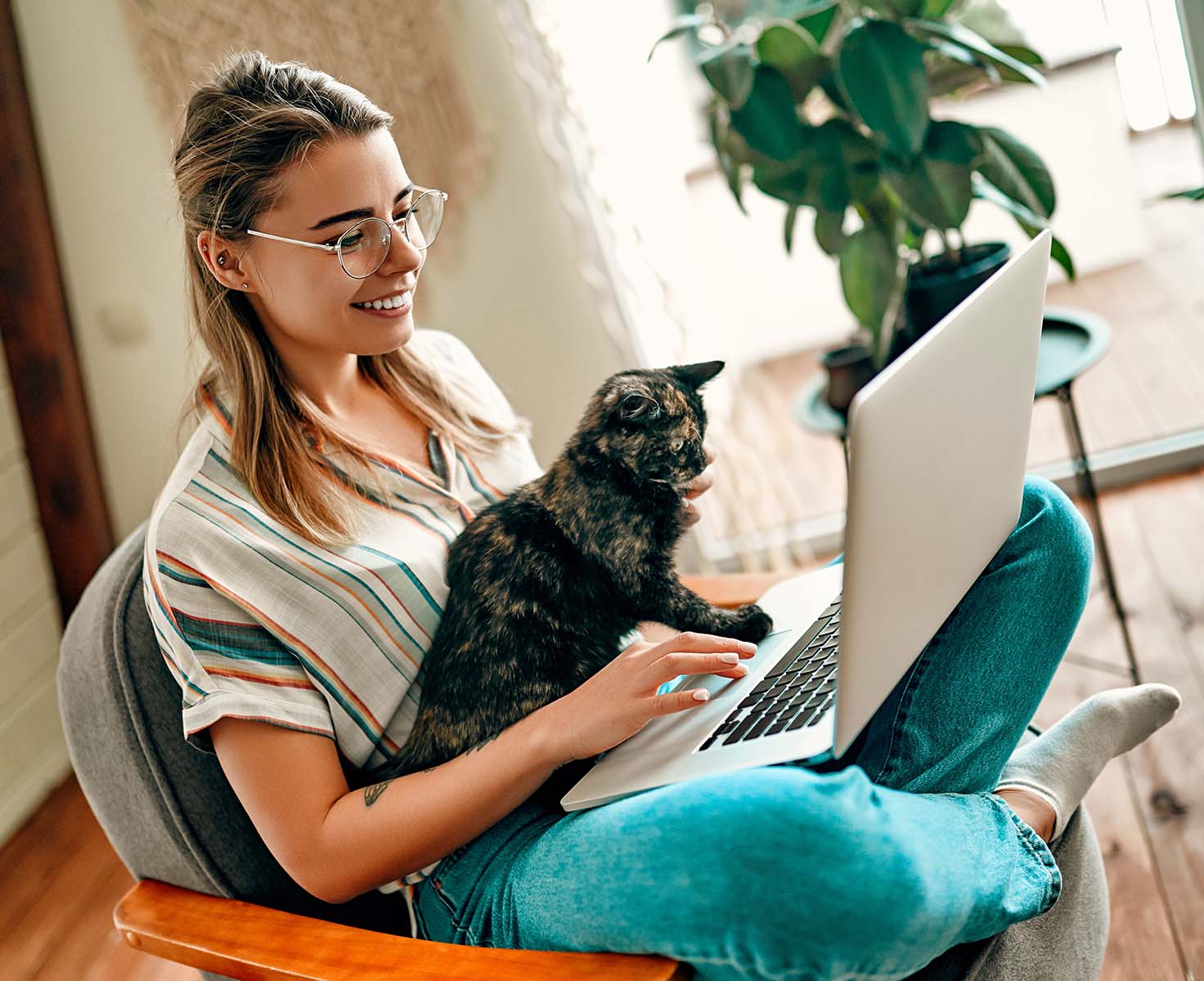 A woman with glasses smiles while working on a laptop with her cat
