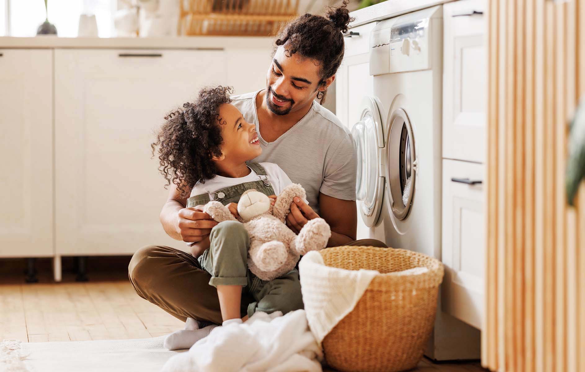 A man and a young child with a teddy bear smile at each other, sitting by a washing machine
