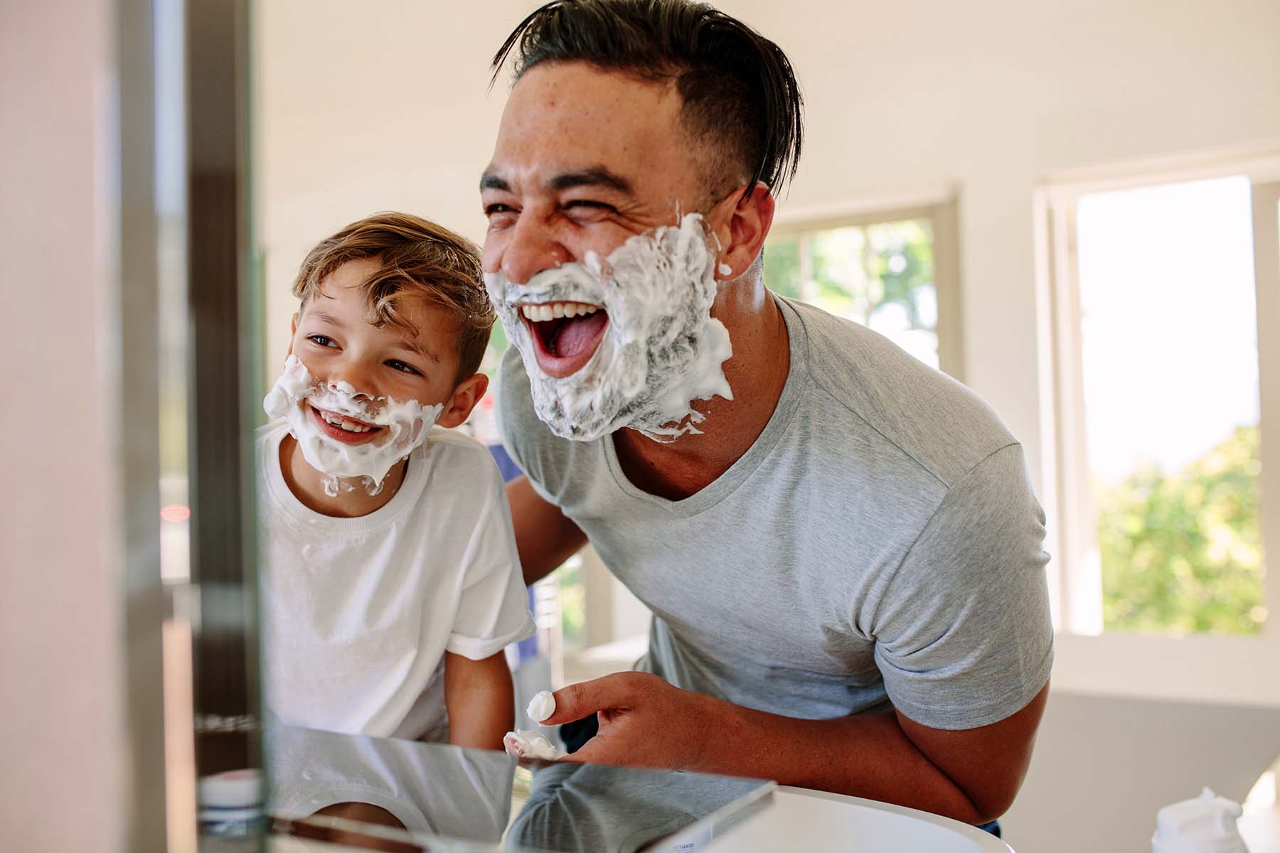 Man and boy with shaving cream on their faces laughing in bathroom.