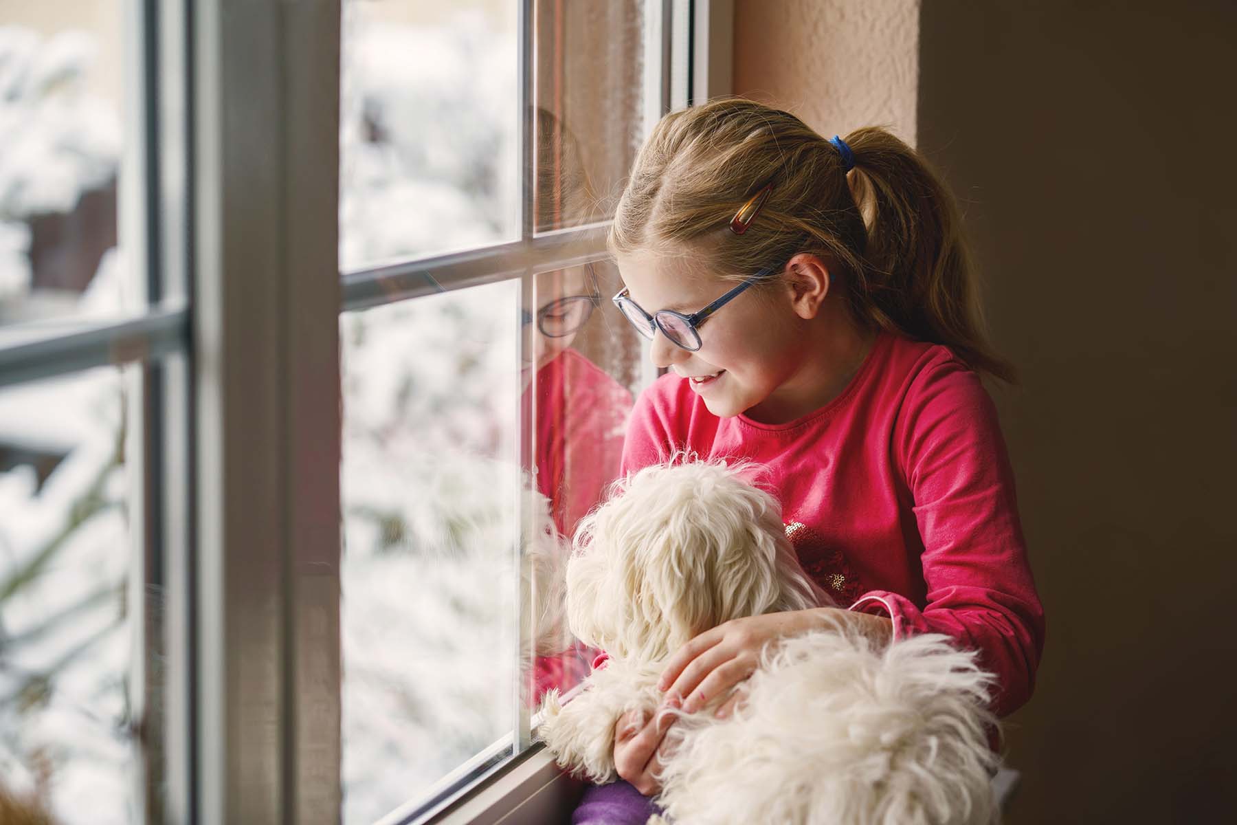 Girl with glasses holding a dog, looking out a snowy window.