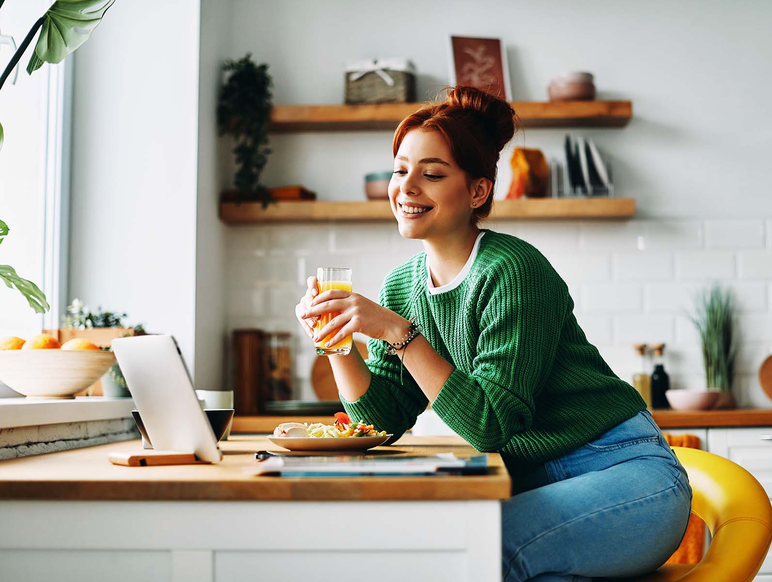 A smiling woman with a drink at a kitchen counter with a laptop.
